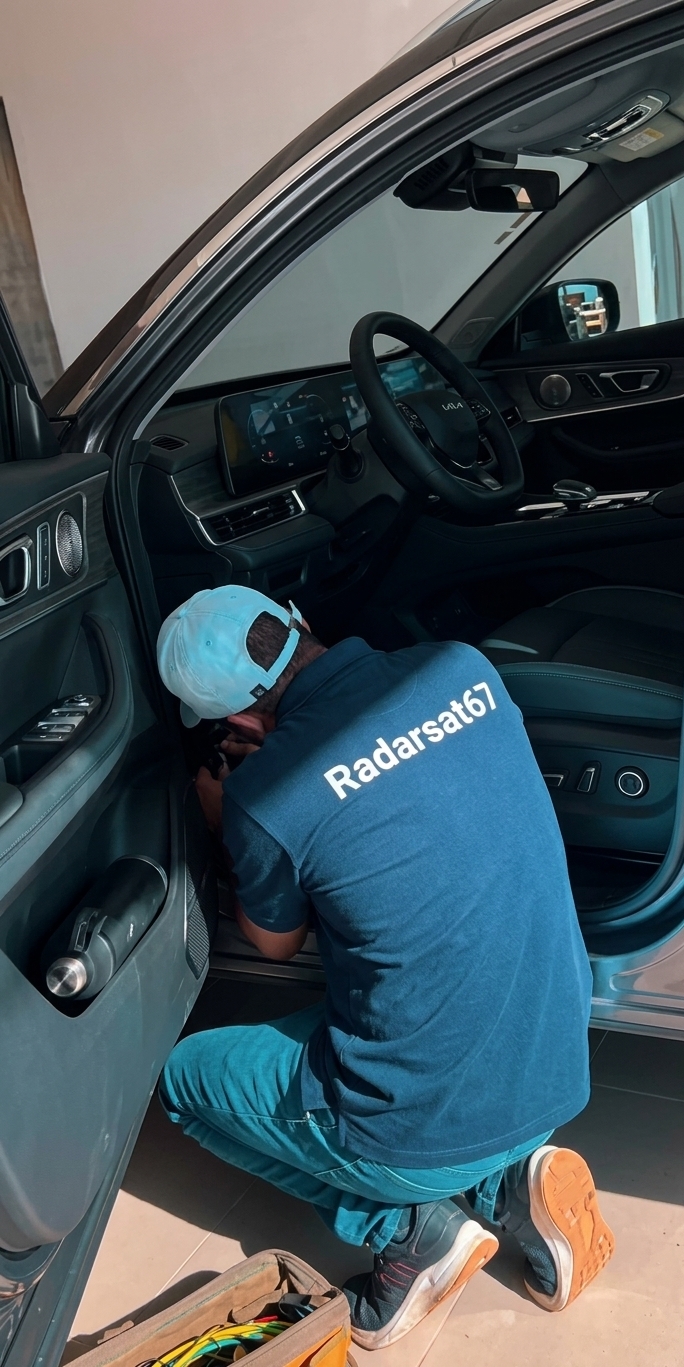 Homem de boné azul e camisa azul com o logo 'Radarsat67' nas costas agachado trabalhando no interior de um car