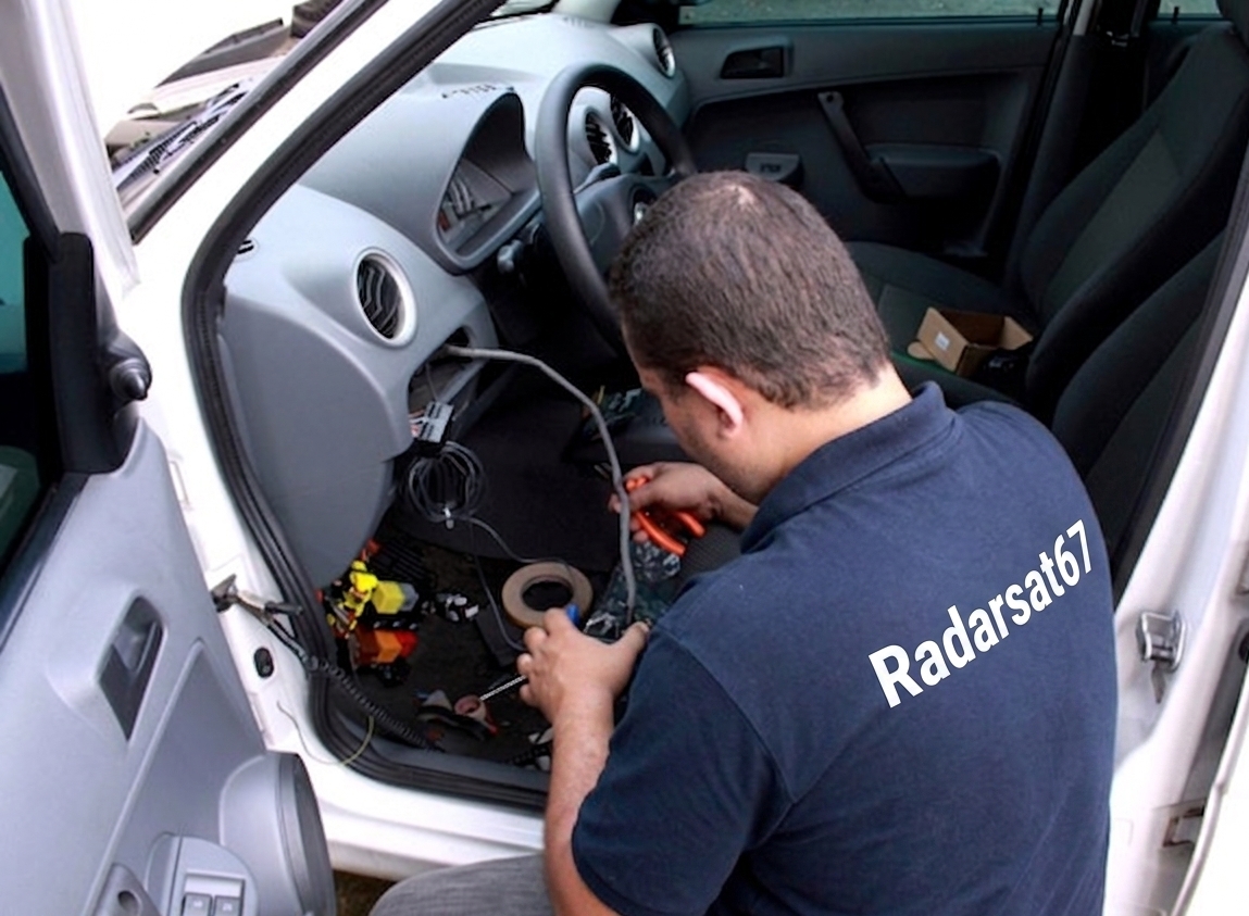 Homem com fone de ouvido instalando equipamento eletrônico no painel de um carro branco, com ferramentas e fio