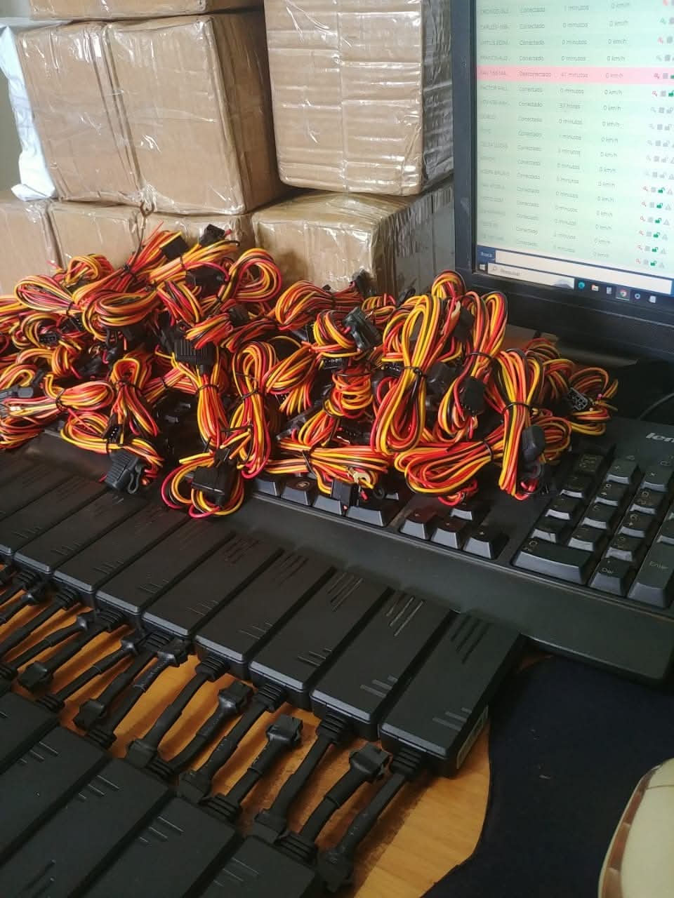 pilha de rastreadores GPS pretos com cabos amarelos e vermelhos enrolados, ao lado de um monitor exibindo soft